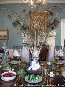 DIY Mardi Gras Centerpiece with peacock feathers in a flower urn sitting on top of a mirrored cast iron tray. A feathered Mardi Gras mask and beads lay at the base of the flower urn